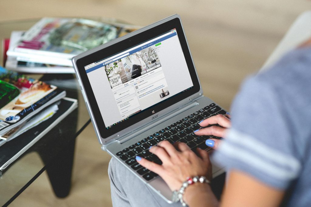 A business owner working on an ad on their laptop after Meta's Andromeda app. Their shoulder, hands, and knees are visible as the laptop sits on their legs, with Facebook visible on the screen.