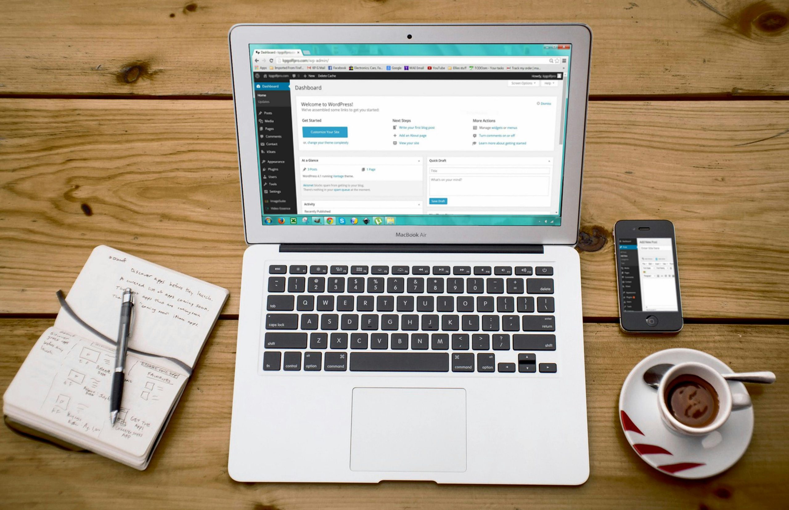 A laptop, notebook, and cup of coffee sitting on a wooden table. The laptop has wordpress visible on the screen.